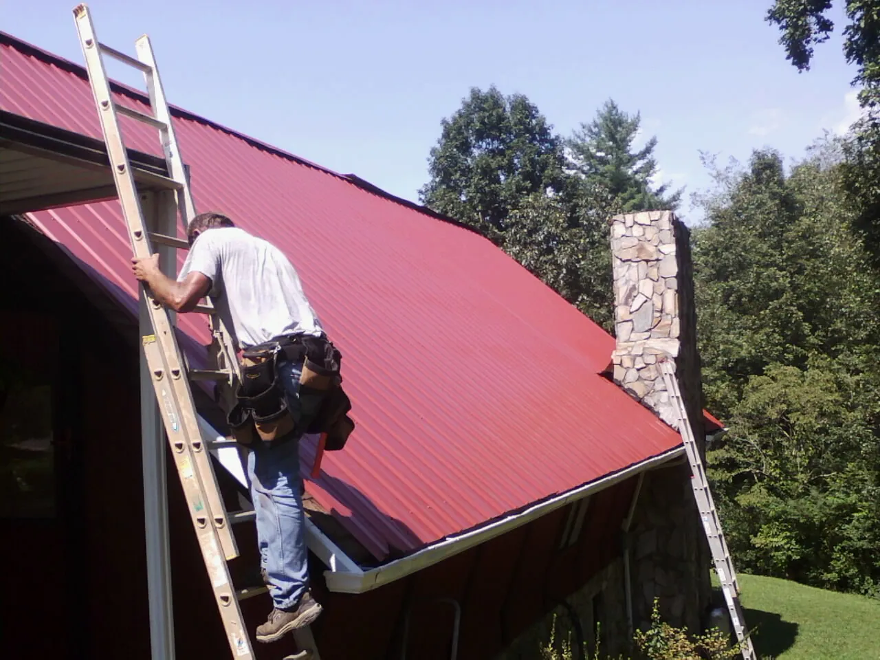 Roofer on a ladder beside a red metal roof and stone chimney — Soffit & Fascia Repair by Roofing Brothers in Arkansas City